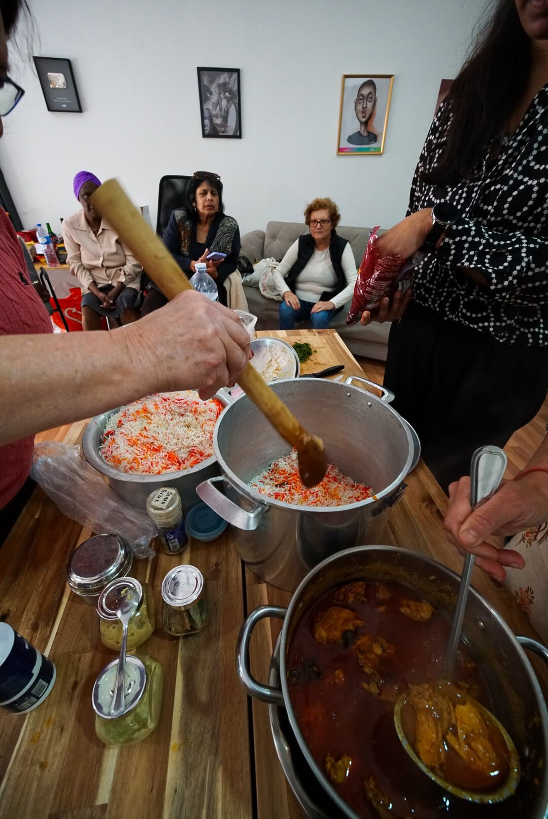 Colher de pau a mexer arroz numa panela, com senhoras sentadas ao fundo